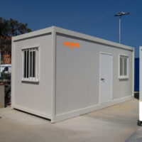 Grey portable office container with a white door and barred windows, branded with an orange HOMS Rentals logo, on a construction site