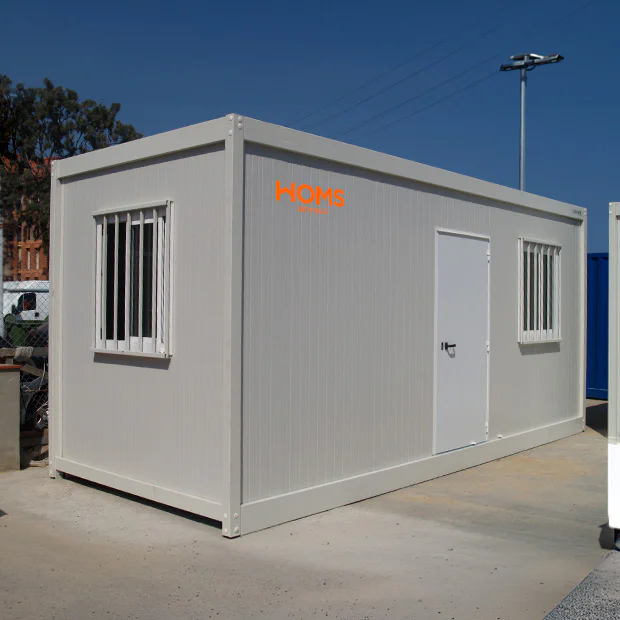 Grey portable office container with a white door and barred windows, branded with an orange HOMS Rentals logo, on a construction site