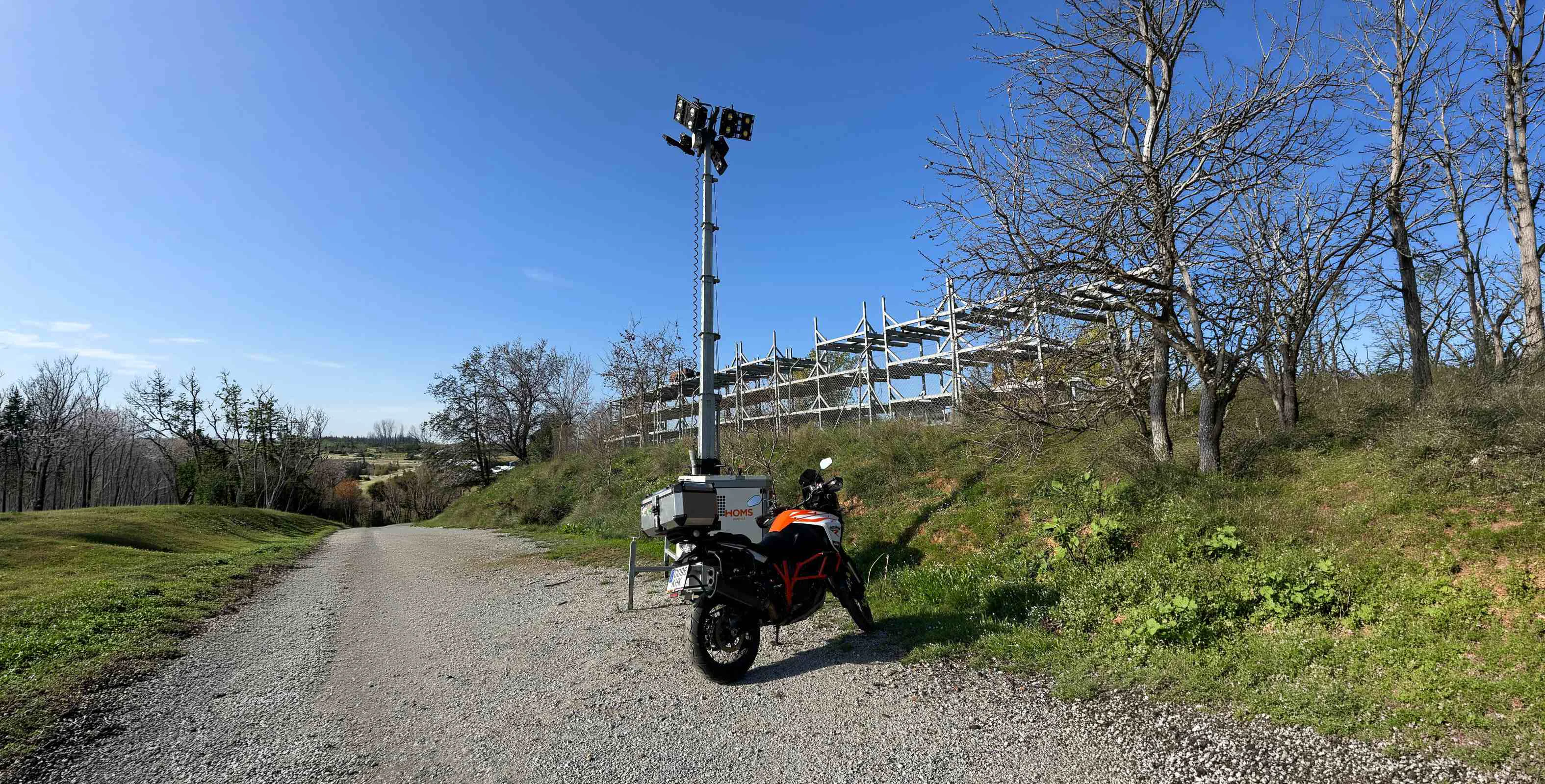 Motorcycle with panniers parked on a gravel road beside a tall floodlight tower and a metal scaffold on a grassy hillside under a blue sky.