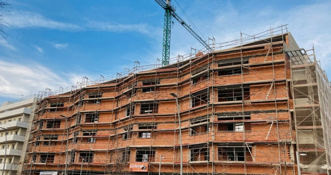 Brick apartment building under construction with metal scaffolding and a tall crane against a blue sky.