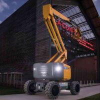 Yellow Haulotte boom lift parked beside a dark building with a red neon sign and a worker basket at its end, night scene.