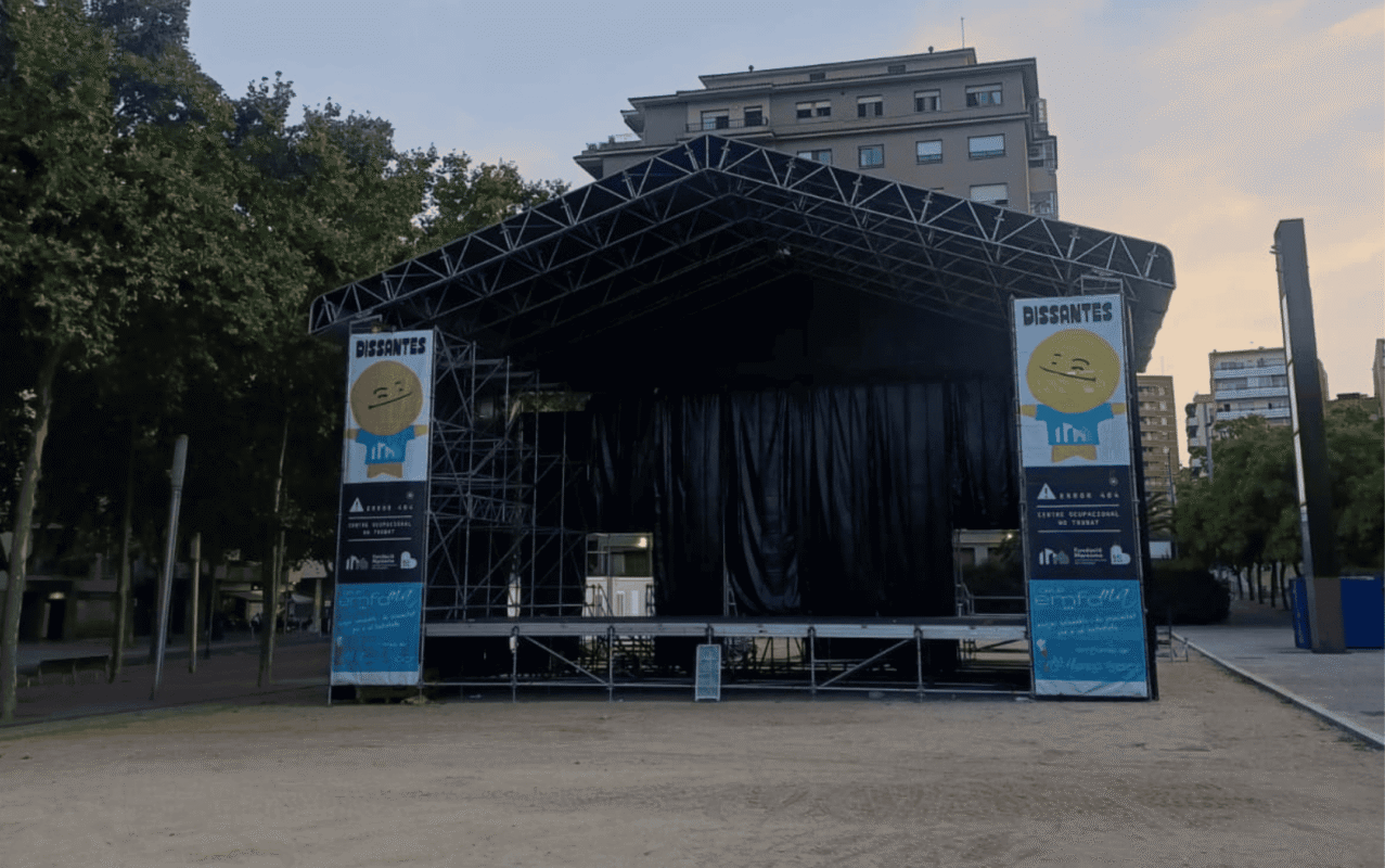Outdoor stage with a black curtain and metal truss roof, flanked by blue banners showing a yellow smiling cartoon figure.