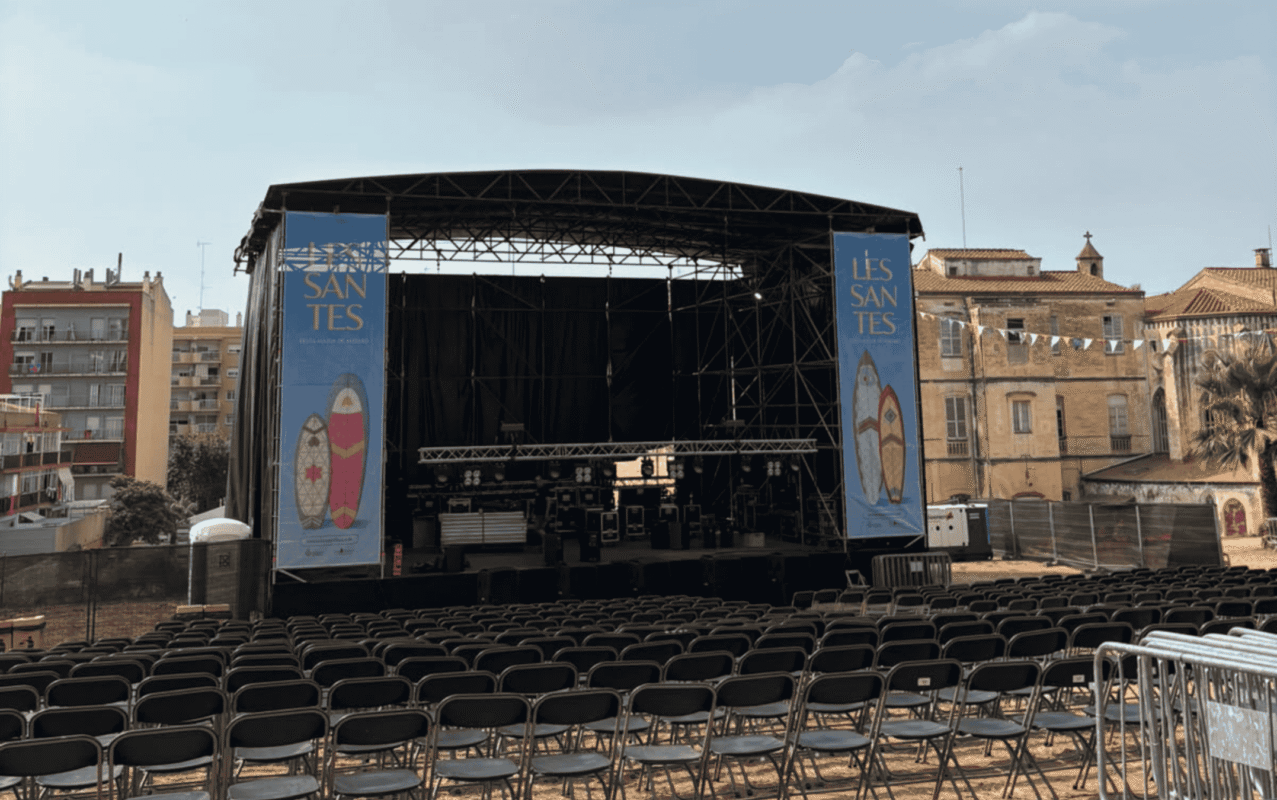 Outdoor festival stage with a metal scaffold, black backdrop, and blue banners on either side, in front of rows of empty chairs in a plaza.