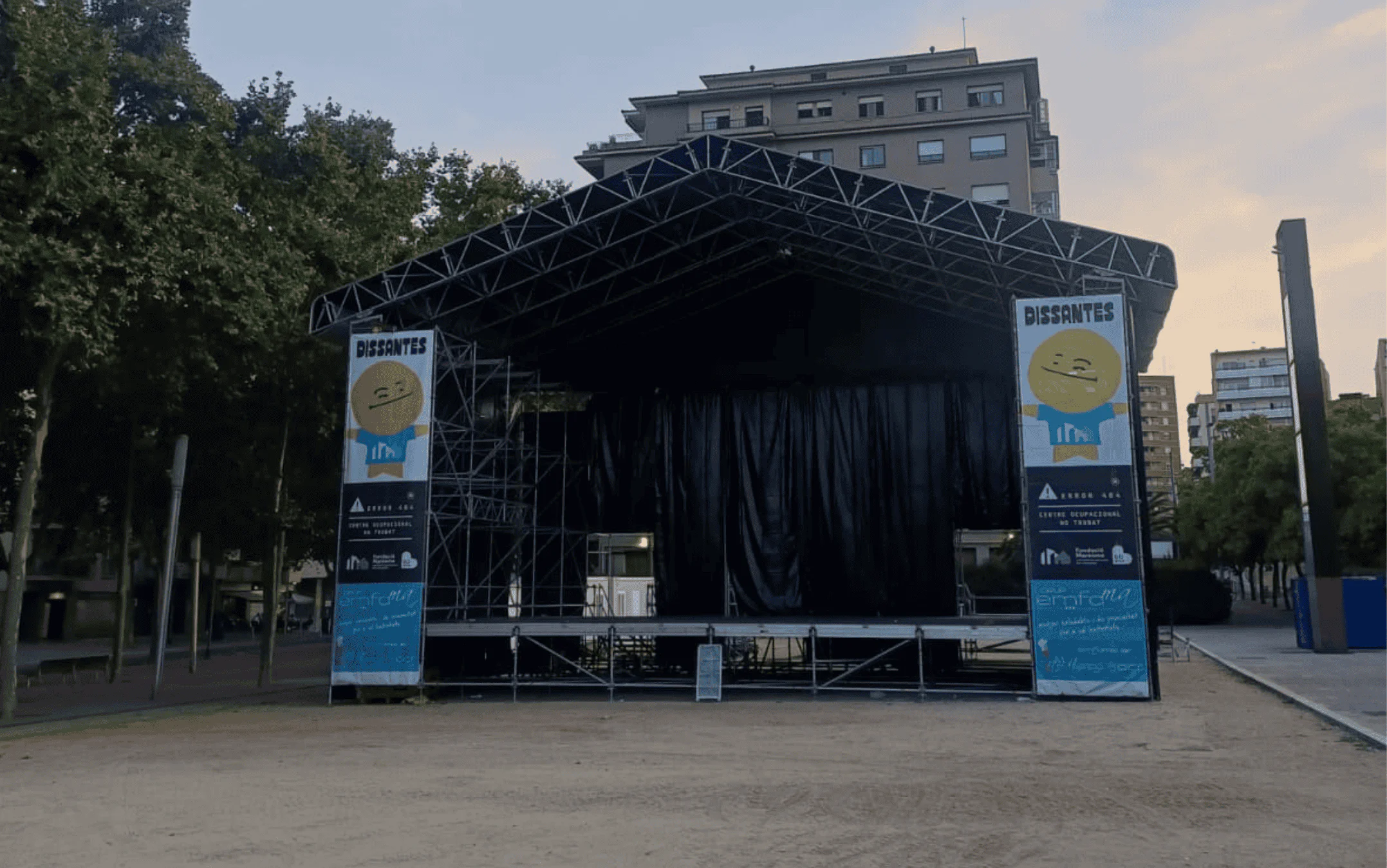 Outdoor stage with black curtains and metal truss, banners featuring a yellow smiley mascot, in a city park at dusk.