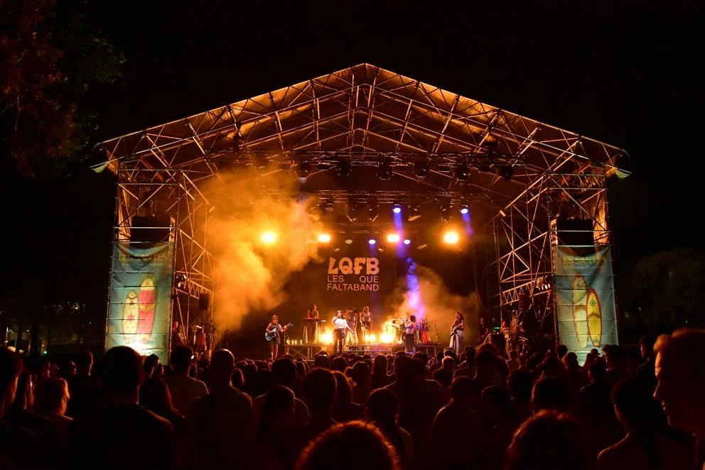 Outdoor nighttime concert: a live band on a large illuminated stage with orange smoke and yellow lights, crowd watching from the foreground.