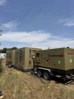 Row of large beige generators on a trailer, connected by cables in a field under a clear blue sky. (Kiloenergia branding visible)