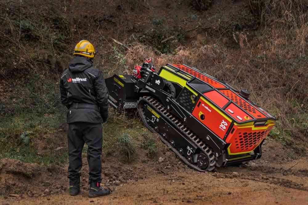 Worker in protective black suit and yellow helmet beside a red and black tracked industrial machine on a dirt slope, inspecting controls.