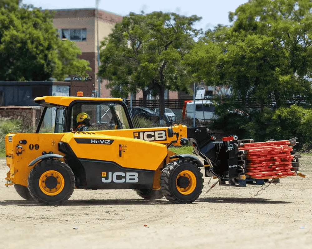 Yellow JCB telehandler on a dirt lot carrying a bundle of red pipes with a clamp attachment, trees in the background.