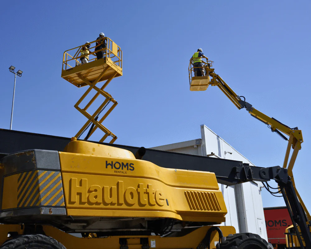 Two workers in safety gear stand on a yellow boom lift platform raised high above a construction site against a clear blue sky. Aerial work platform with workers performing tasks on an elevated basket (boom lift). Bright yellow Haulotte rental boom lift with workers in the basket on a sunny day. Elevated work platform in use: workers on a yellow hydraulic boom lift performing tasks. Wide shot of a yellow hydraulic boom lift beside a building under a clear blue sky.