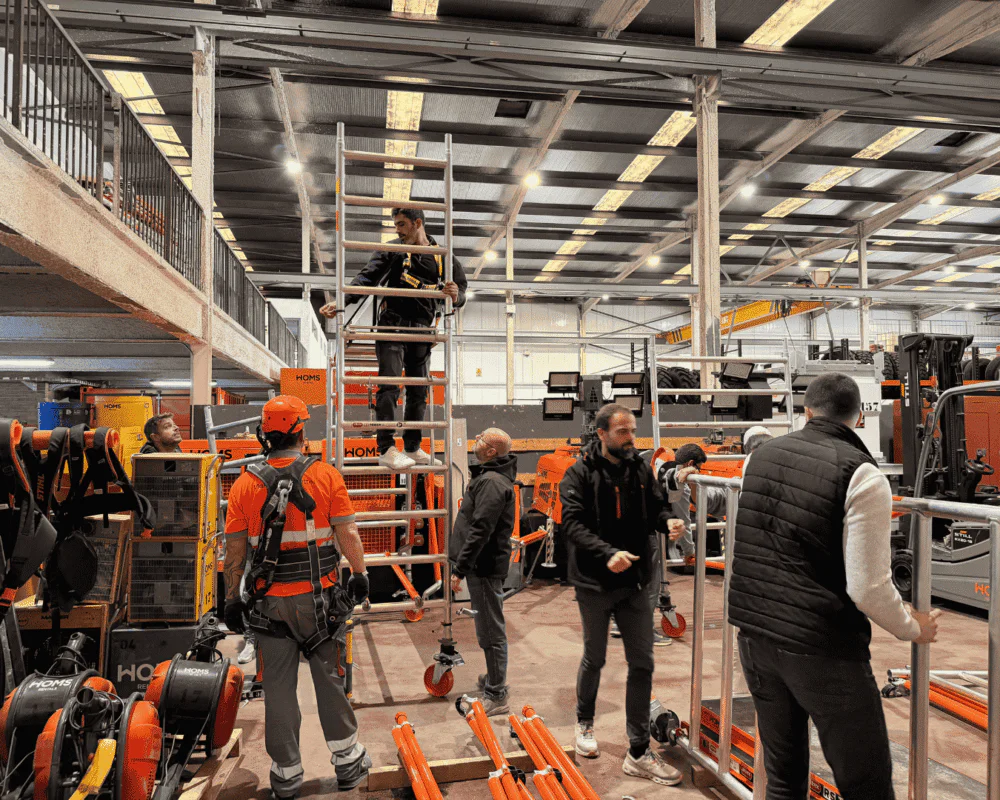 Warehouse workshop with workers assembling orange scaffolding; a man stands on a tall ladder while others assist nearby, all wearing safety gear and helmets.