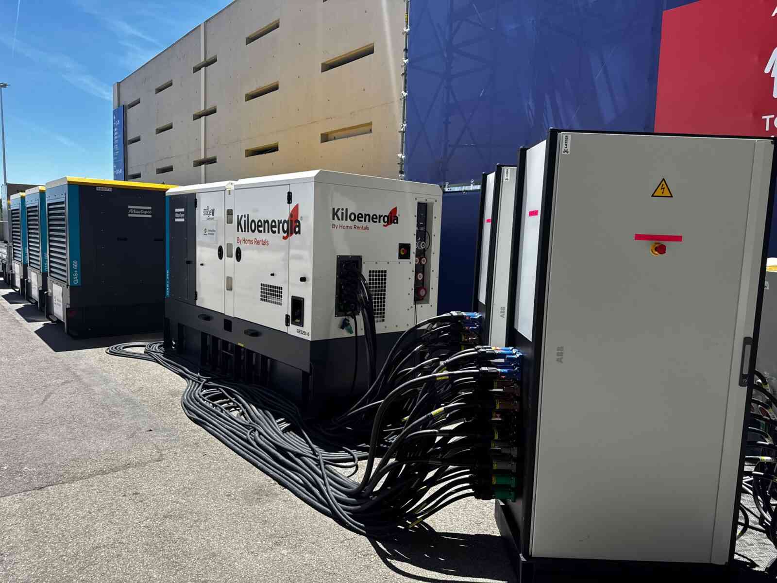 Line of large industrial generators and electrical cabinets outdoors, with thick black cables coiled on the ground and a blue sky above.