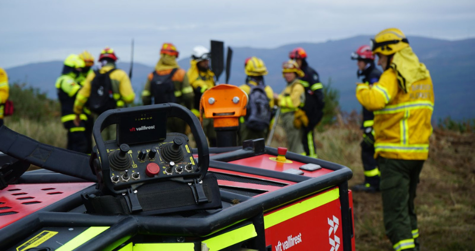 Firefighters in yellow gear gather around a red rescue vehicle with equipment on top, on a hill with mountains in the background.