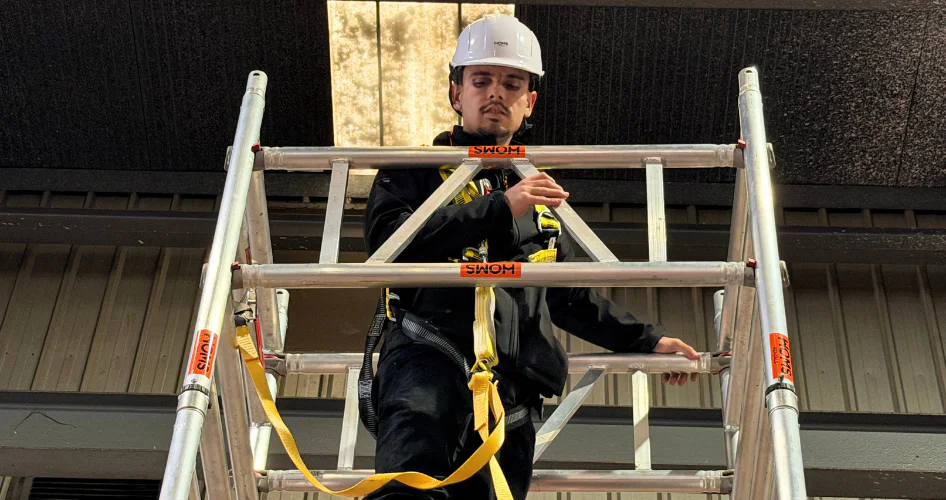 Worker in a white hard hat and black safety gear on a metal scaffold, adjusting a yellow fall-protection strap indoors.